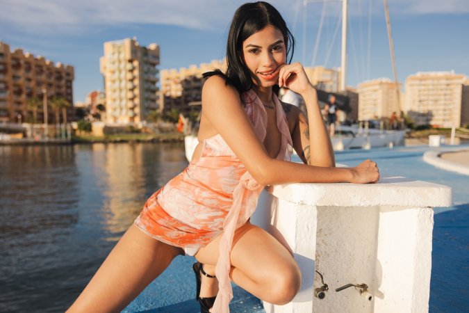 Sofia sits bare-legged on a blue marina dock in a sheer swimsuit, teasing with no underwear as boats drift behind her — Sofia #20