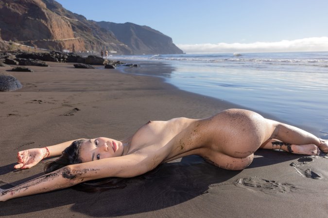 Sky Moon bending over rainbow towel on black sand beach, sunlight warming her naked curves — Sky Moon #11