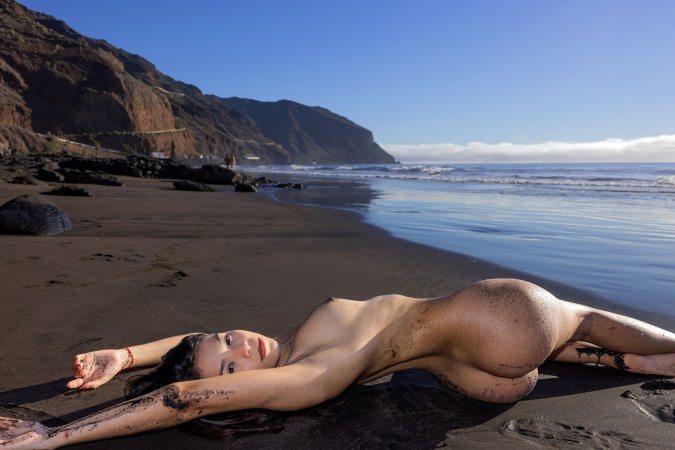 Sky Moon bending over rainbow towel on black sand beach, sunlight warming her naked curves — Sky Moon #12