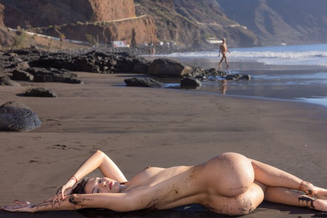 Sky Moon bending over rainbow towel on black sand beach, sunlight warming her naked curves — Sky Moon #15