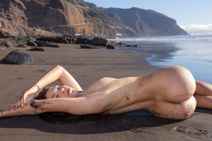 Sky Moon bending over rainbow towel on black sand beach, sunlight warming her naked curves — Sky Moon #16