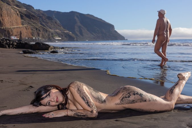 Sky Moon bending over rainbow towel on black sand beach, sunlight warming her naked curves — Sky Moon #17