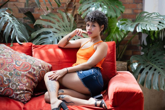 Lilith Baph poses solo on a red couch in orange top and denim shorts, tattooed legs spread for the lens — Lilith Baph #1
