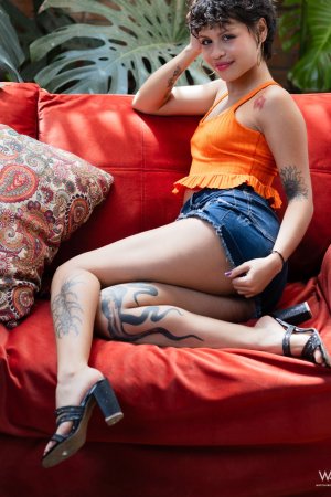 Lilith Baph poses solo on a red couch in orange top and denim shorts, tattooed legs spread for the lens — Lilith Baph #2