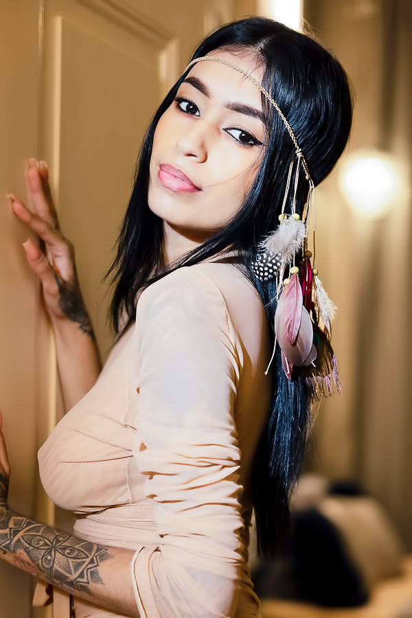 Alexa Belluci poses in beige dress with feather earrings, tattoos visible, indoor chandelier glow