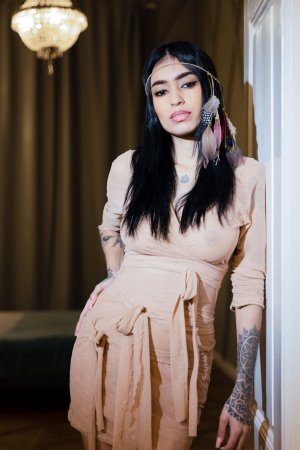 Alexa Belluci poses in beige dress with feather earrings, tattoos visible, indoor chandelier glow — Alexa Belluci #1