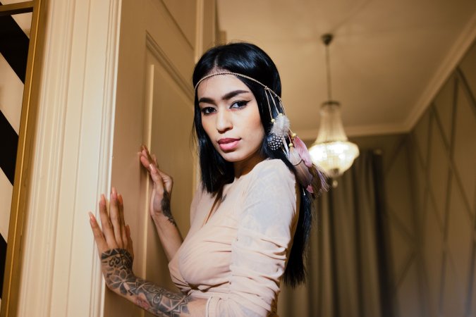 Alexa Belluci poses in beige dress with feather earrings, tattoos visible, indoor chandelier glow — Alexa Belluci #4