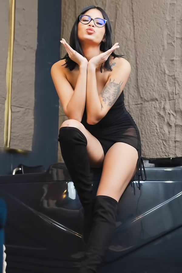 Sofia poses in black lingerie and thigh high boots beside a velvet bed, tattooed arm raised, orchids nearby