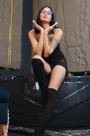 Sofia poses in black lingerie and thigh high boots beside a velvet bed, tattooed arm raised, orchids nearby — Sofia #4