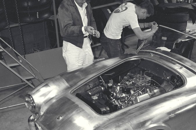 Nika leans against a vintage AC Cobra in the garage heat, her gaze locked on the racetrack ahead — Nika #1