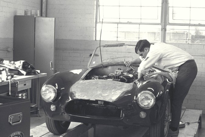 Nika leans against a vintage AC Cobra in the garage heat, her gaze locked on the racetrack ahead — Nika #2