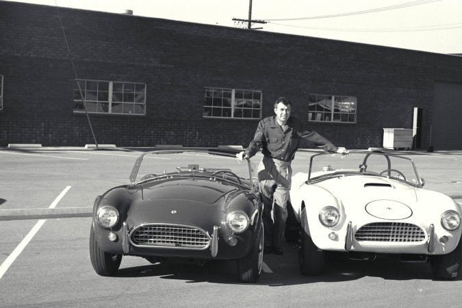 Nika leans against a vintage AC Cobra in the garage heat, her gaze locked on the racetrack ahead — Nika #3