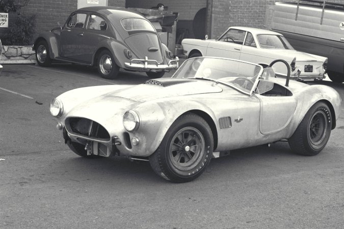 Nika leans against a vintage AC Cobra in the garage heat, her gaze locked on the racetrack ahead — Nika #4