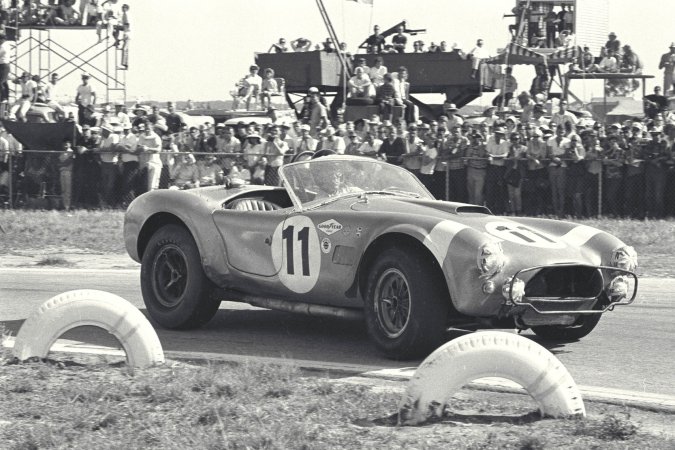 Nika leans against a vintage AC Cobra in the garage heat, her gaze locked on the racetrack ahead — Nika #5