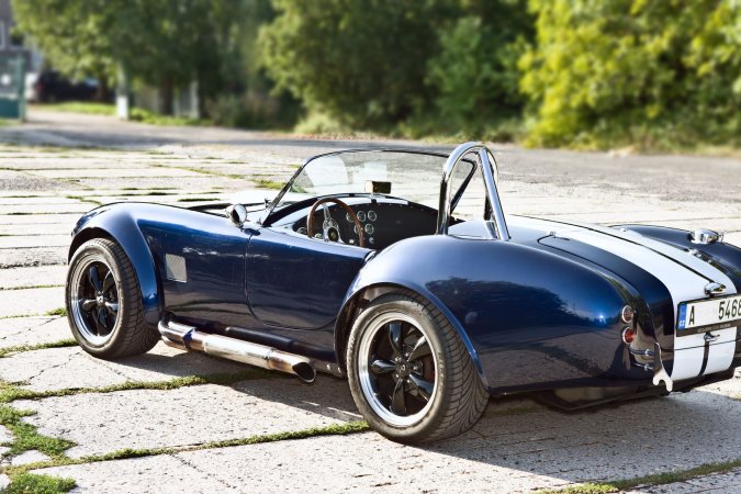 Nika leans against a vintage AC Cobra in the garage heat, her gaze locked on the racetrack ahead — Nika #9