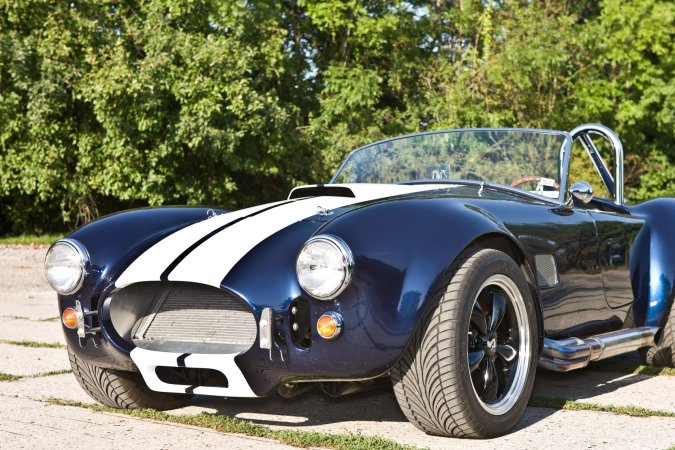 Nika leans against a vintage AC Cobra in the garage heat, her gaze locked on the racetrack ahead — Nika #11