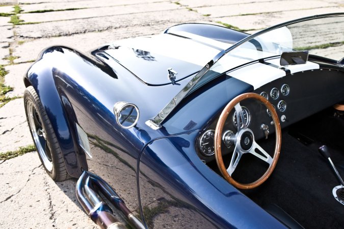 Nika leans against a vintage AC Cobra in the garage heat, her gaze locked on the racetrack ahead — Nika #12