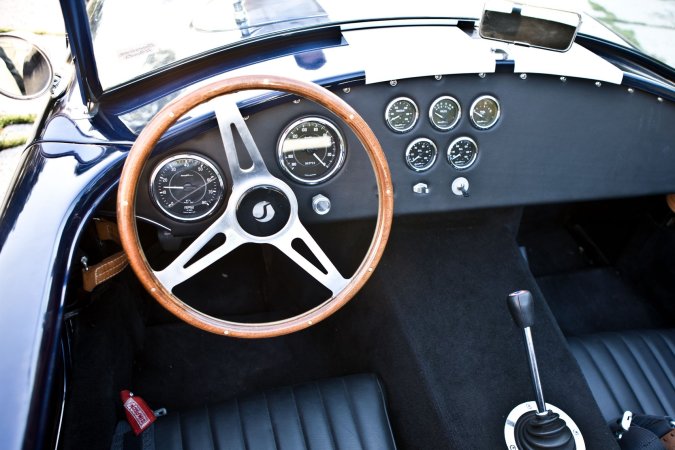 Nika leans against a vintage AC Cobra in the garage heat, her gaze locked on the racetrack ahead — Nika #13