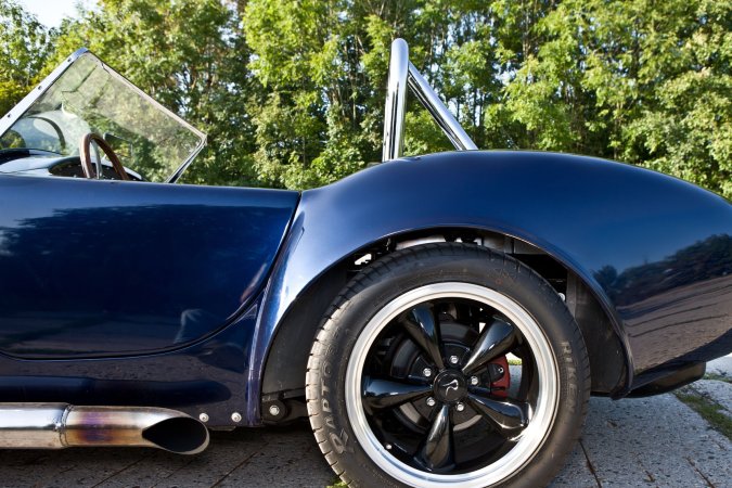 Nika leans against a vintage AC Cobra in the garage heat, her gaze locked on the racetrack ahead — Nika #14