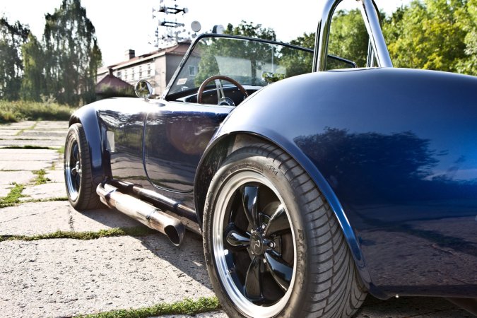 Nika leans against a vintage AC Cobra in the garage heat, her gaze locked on the racetrack ahead — Nika #15